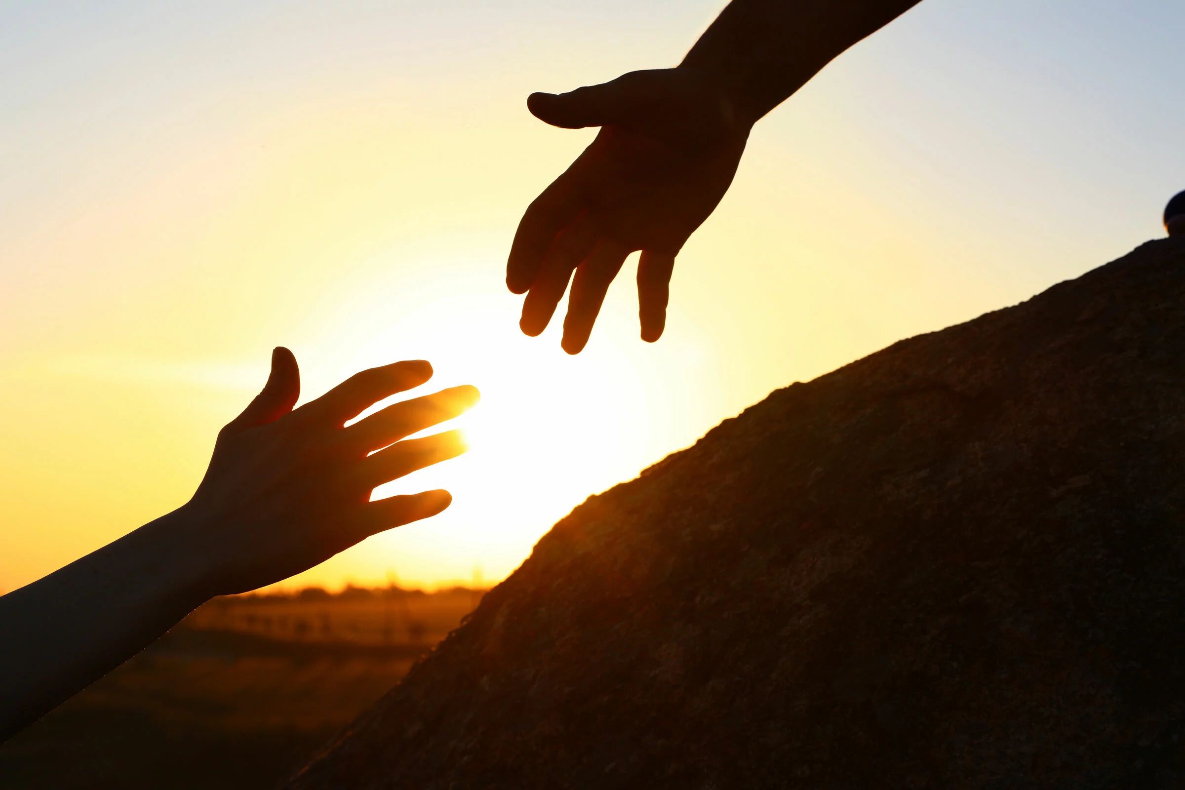 Zwei Hände reichen sich im Sonnenuntergang über einem Hügel die Hand. Die Szene vermittelt Unterstützung, Trost und Zusammenhalt in einer Trauersituation.